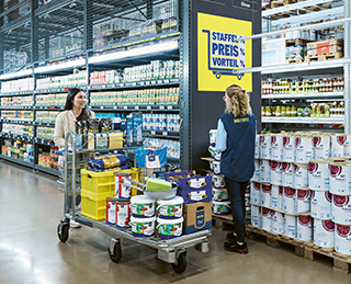 Eine Frau die einen vollen Einkaufswagen vor sich herschiebt und eine METRO Mitarbeiterin anlächelt (Foto)