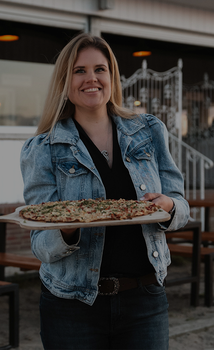 Eine lachende Frau, die einen Flammkuchen auf einem Brett hält. (Foto)