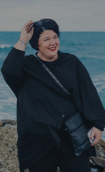 Eine lachende Frau mit Handtasche, Sonnenbrille und Smartphone am Meer. (Foto)