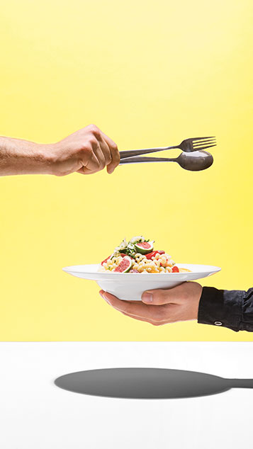 Bunte Mahlzeit präsentiert in einem weißen Teller, getragen von einer Hand; Zweite Hand Besteck haltend ragt darüber in das Bild (Foto)